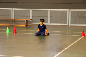 Fotografia de um menino sentado no chão da quadra de esportes com uma venda no olho. A atividade é a goalball. O menino está com o uniforem azul marinho do Colégio Miguel de Cervantes.