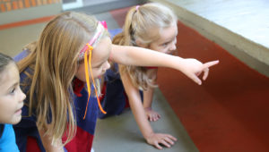 3 alunas do Infantil 5 estão agachadas, olhando para a parte de baixo de um banco do pátio. A aluna do meio está apontando o dedo indicador para sua frente.
